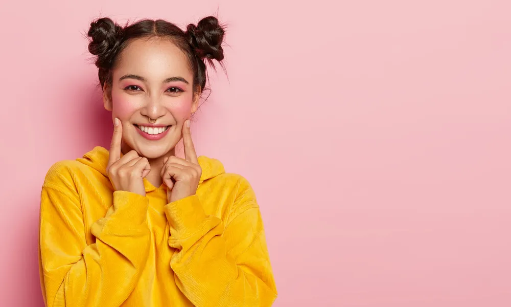 beautiful-brunette-woman-with-two-buns-points-index-fingers-cheeks-dressed-casual-corduoy-yellow-sweatshirt-wears-bright-pinup-makeup