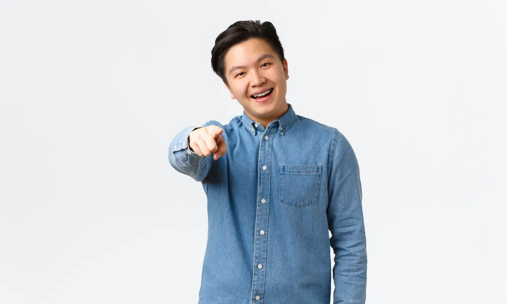 smiling-handsome-asian-guy-with-braces-making-choice-pointing-finger-camera-choosing-picking-something-found-person-standing-white-background-congratulating-you